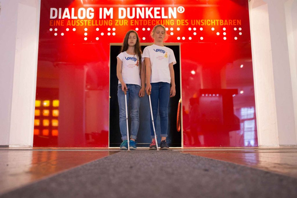 Zwei junge BesucherInnen vor dem Eingang der Ausstellung, mit einem Blindenstock, der den Leitstreifen vor Ihnen berührt.