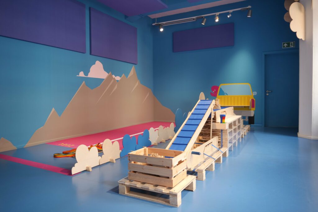 Ein Spielbereich für Kinder mit einer Berg- und Himmelmotiv-Wandmalerei, einem hölzernen Förderband auf blauem Boden.