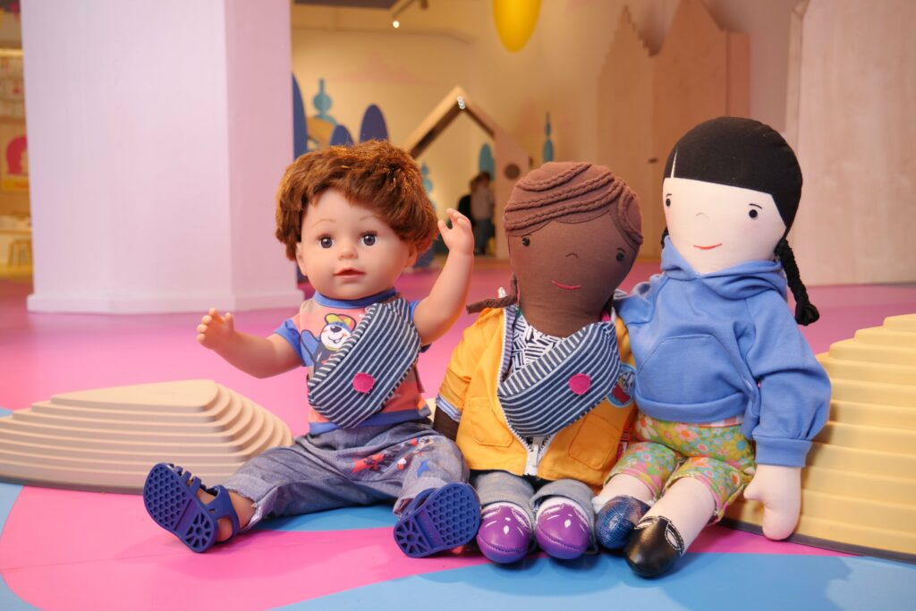 Drei verschiedene Spielzeugpuppen auf einer bunten Matte, die Vielfalt und Inklusion darstellen, mit einer Puppe, die einen europäischen Teint hat, einer Puppe mit einem afrikanischen Teint und einer Puppe mit einem asiatischen Teint.