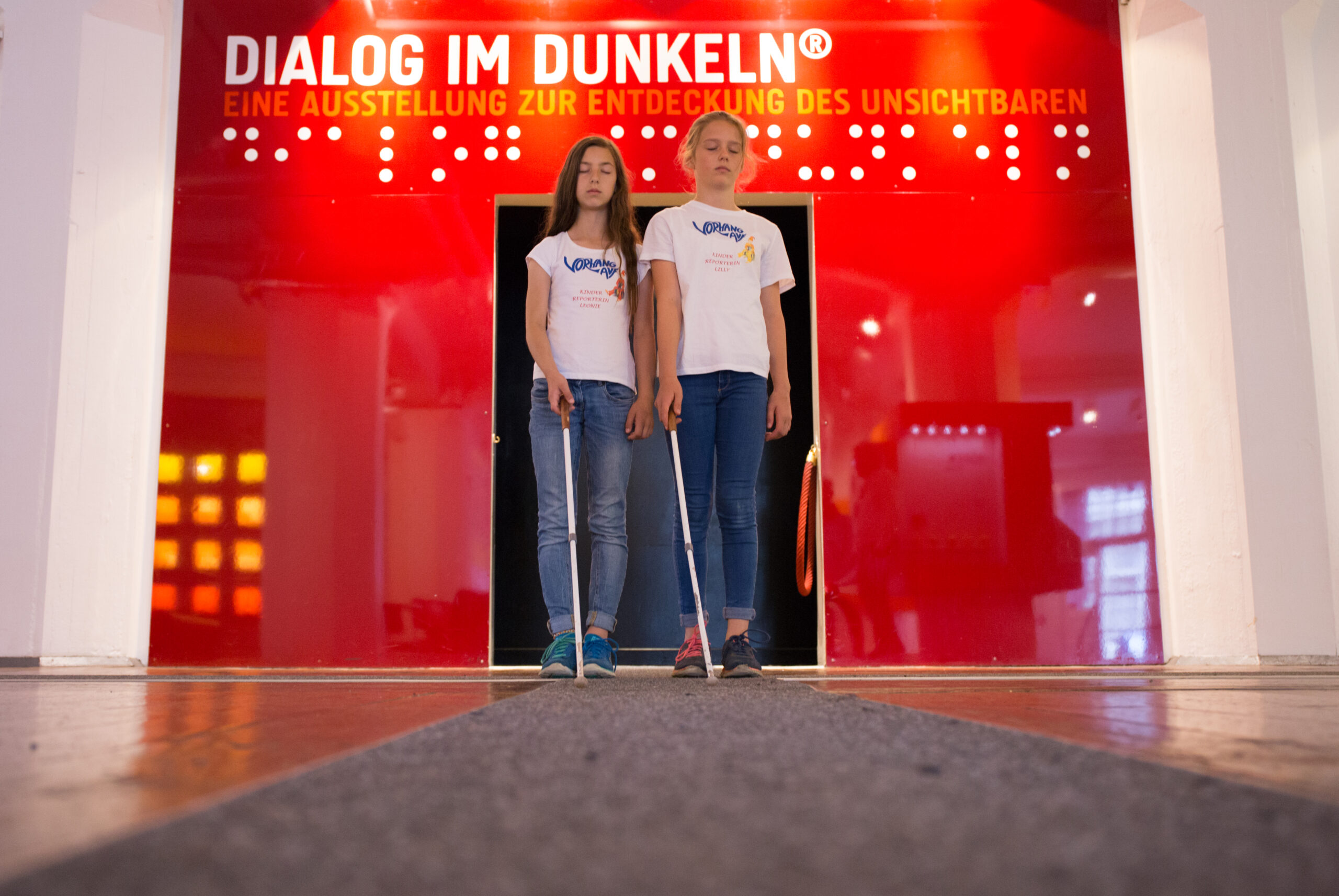 Zwei Schülerinnen stehen, jeweils mit einem weißen Langstock in der Hand, vor dem Eingang zum Dialog im Dunkeln.