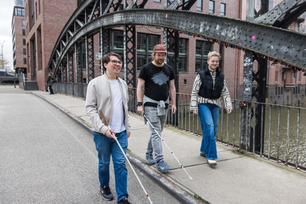 3 Mitarbeitende des Dialoghaus Hamburg laufen in der Speicherstadt entland. Zwei von ihnen nutzen einen Leitstock zur Orientierung.