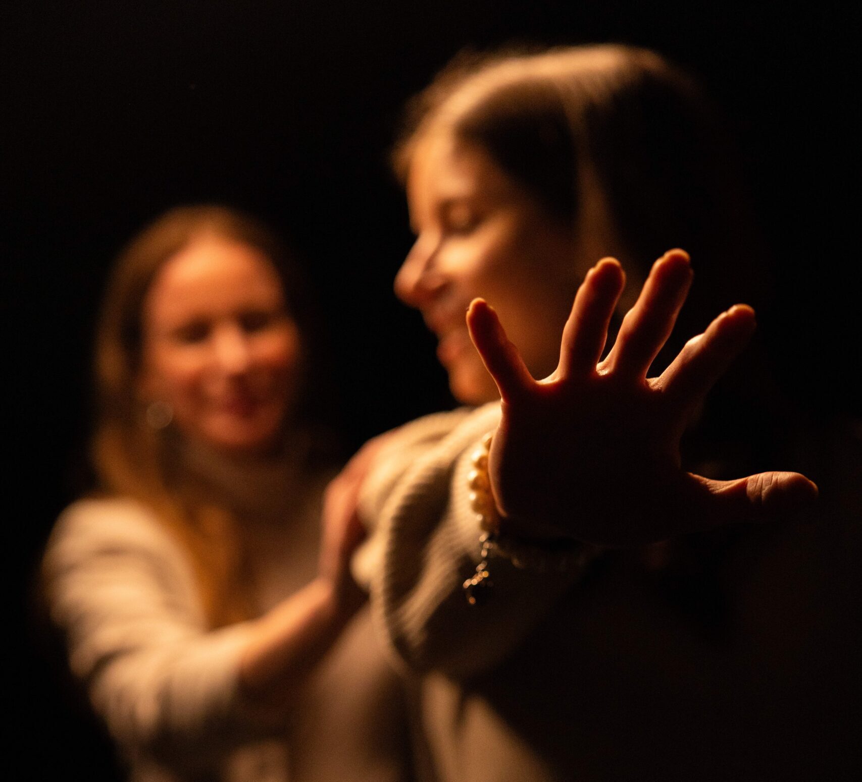 Eine Frau streckt in einer dunklen Umgebung suchend die Hand nach vorne, während eine zweite Frau hinter ihr steht und ihre Hand auf die Schulter der Frau vor ihr legt.