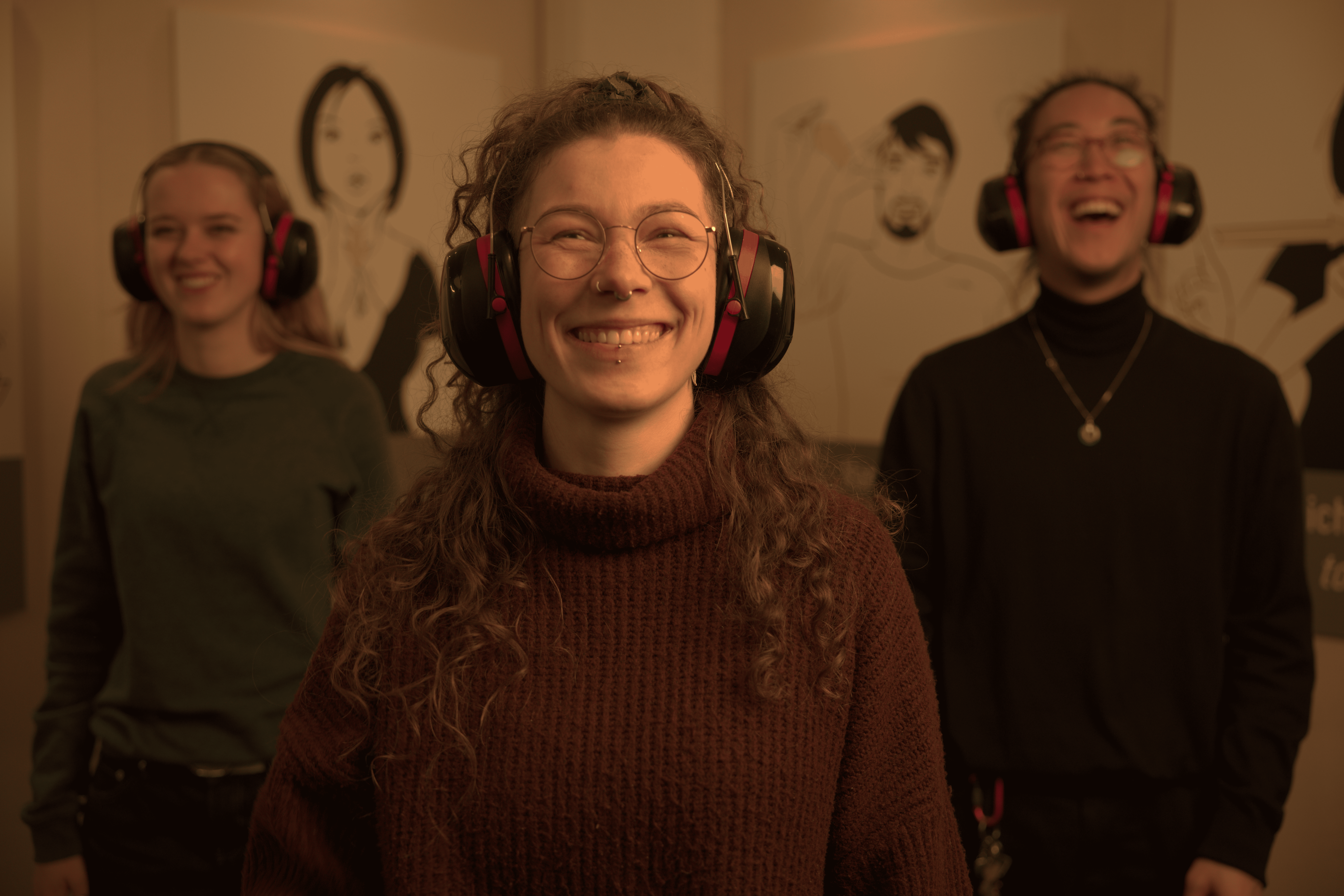 Eine lächelnde Frau mit schalldichten Kopfhörern steht im Zentrum des Bilds. Hinter ihr stehen links und rechts leicht versetzt zwei weitere Personen mit Kopfhörern.