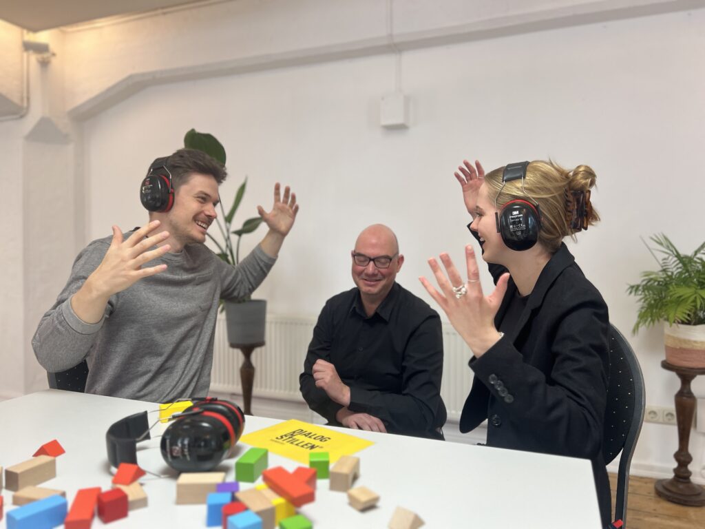 Ein gehörloser Guide unterhält sich mit zwei Workshop-Teilnehmenden mithilfe von Gebärden. Die beiden Teilnehmenden haben große schalldichte Kopfhörer über ihren Ohren.