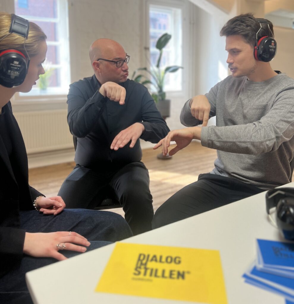 Ein gehörloser Guide unterhält sich mit zwei Workshop-Teilnehmenden mithilfe von Gebärden. Die beiden Teilnehmenden haben große schalldichte Kopfhörer über ihren Ohren.