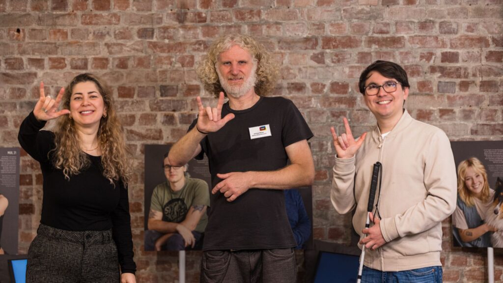 3 Mitarbeitende des Dialoghaus Hamburg stehen im Foyer des Hauses und halten die "I love you"-Gebärde mit jeweils einer Hand in Richtung Kamera. Eine der Personen hält einen Langstock in der anderen Hand.
