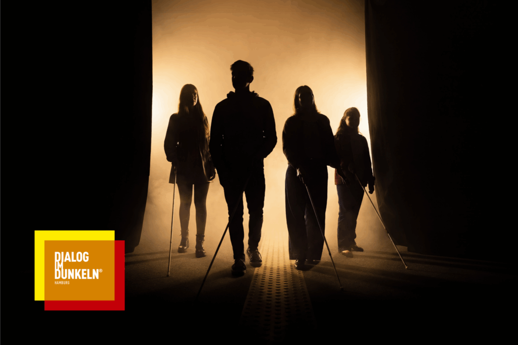 Schüler*innen bewegen sich gemeinsam mit blinden und sehbehinderten Guides durch eine lichtlose Ausstellung. Die Gruppe orientiert sich mit Langstöcken in einem nachgestellten Alltagsraum bei „Dialog im Dunkeln“ im Dialoghaus Hamburg.