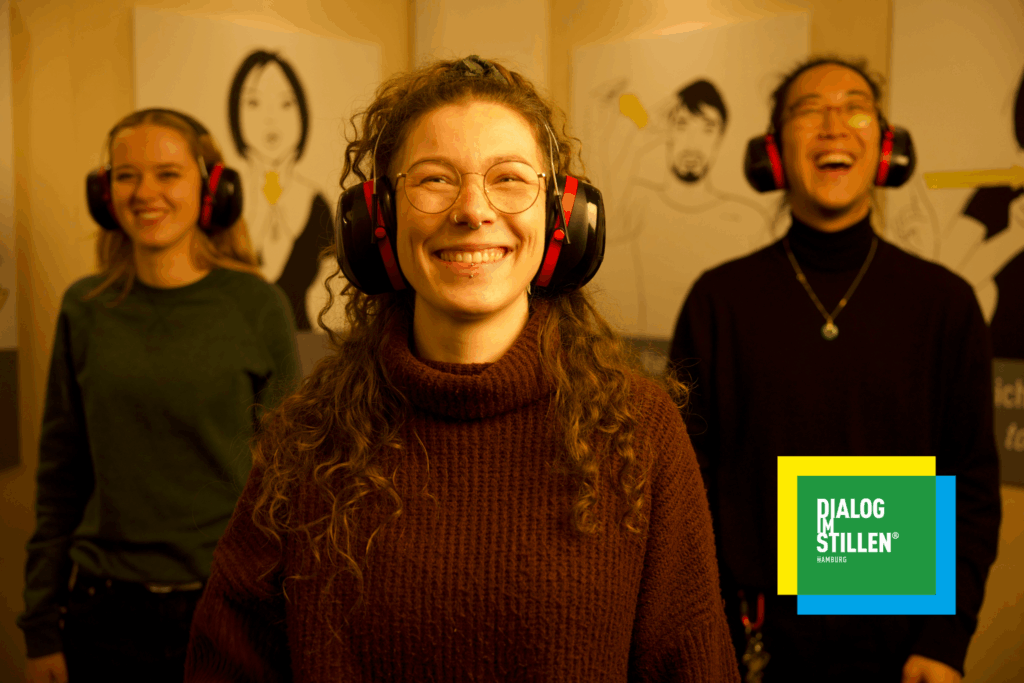 Schüler*innen tragen Gehörschutz und stehen gemeinsam in der Ausstellung „Dialog im Stillen“. Im Vordergrund eine Person mit Kopfhörern, im Hintergrund weitere Teilnehmende – Kommunikation erfolgt über Gestik und Mimik.