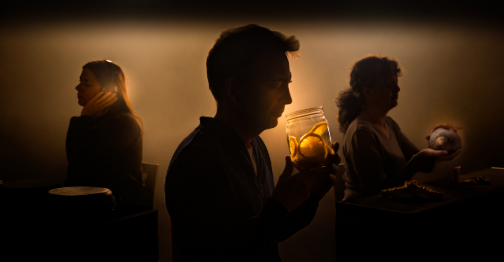 Drei Personen sitzen in einer warm beleuchteten, leicht nebligen Umgebung und konzentrieren sich auf ihre Sinne: Eine Person riecht an einem Glas mit Zitronenscheiben, eine andere ertastet ein Objekt, während eine dritte sich auf Geräusche konzentriert.