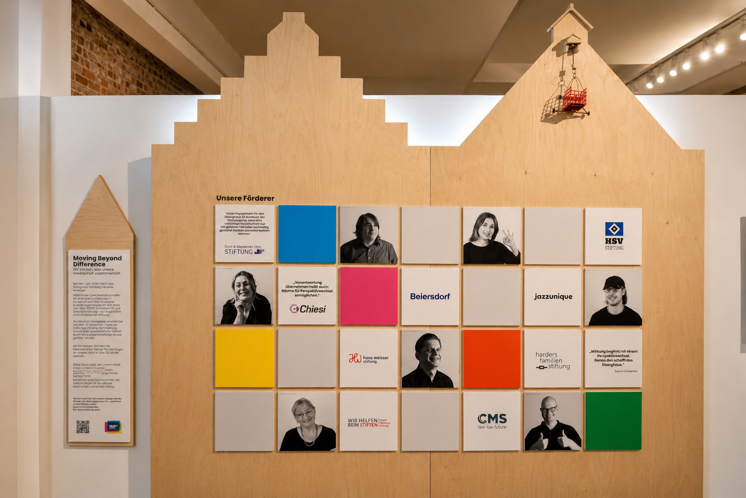 Große Förderwand auf einer holzverkleideten Ausstellungsfläche mit bunten Kacheln, Porträts, Zitaten und Logos von Unterstützer:innen des Dialoghaus Hamburg.