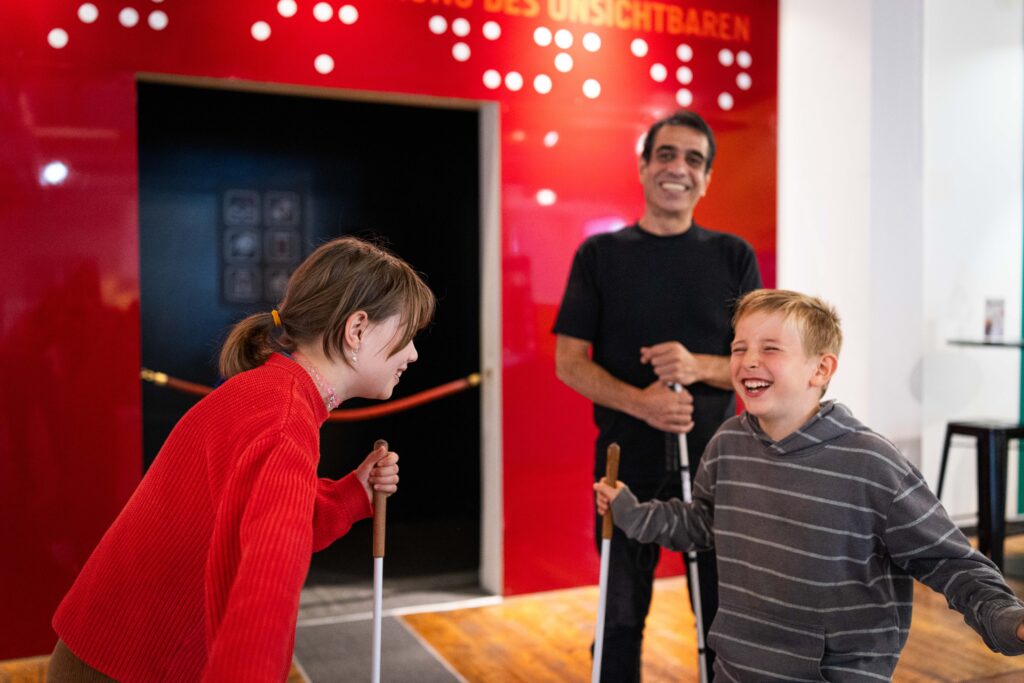 Zwei Kinder lachen mit Langstöcken vor dem Eingang zu Dialog im Dunkeln. Guide Kourosh steht lächelnd dahinter.