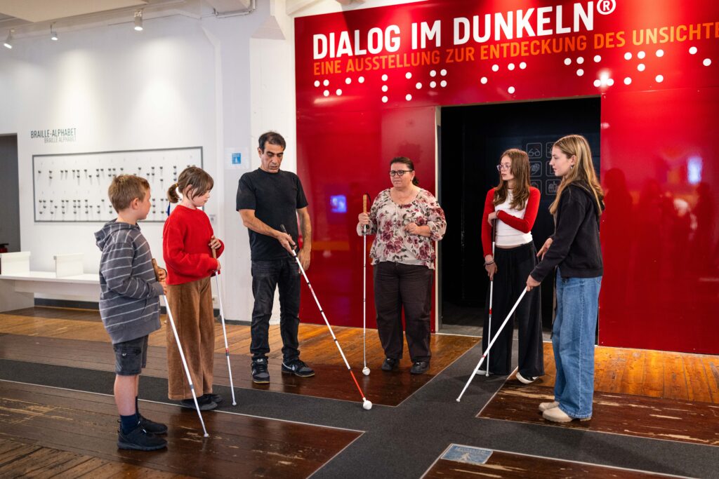 Vier Kinder stehen mit den Guides Kourosh und Silvana mit Langstöcken vor dem Eingang zu Dialog im Dunkeln.