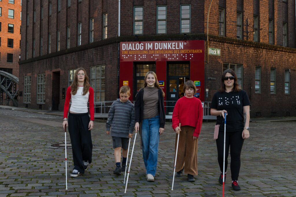 Vier Kinder und Guide Anna stehen mit Langstöcken vor dem Eingang zu Dialog im Dunkeln in der Hamburger Speicherstadt.